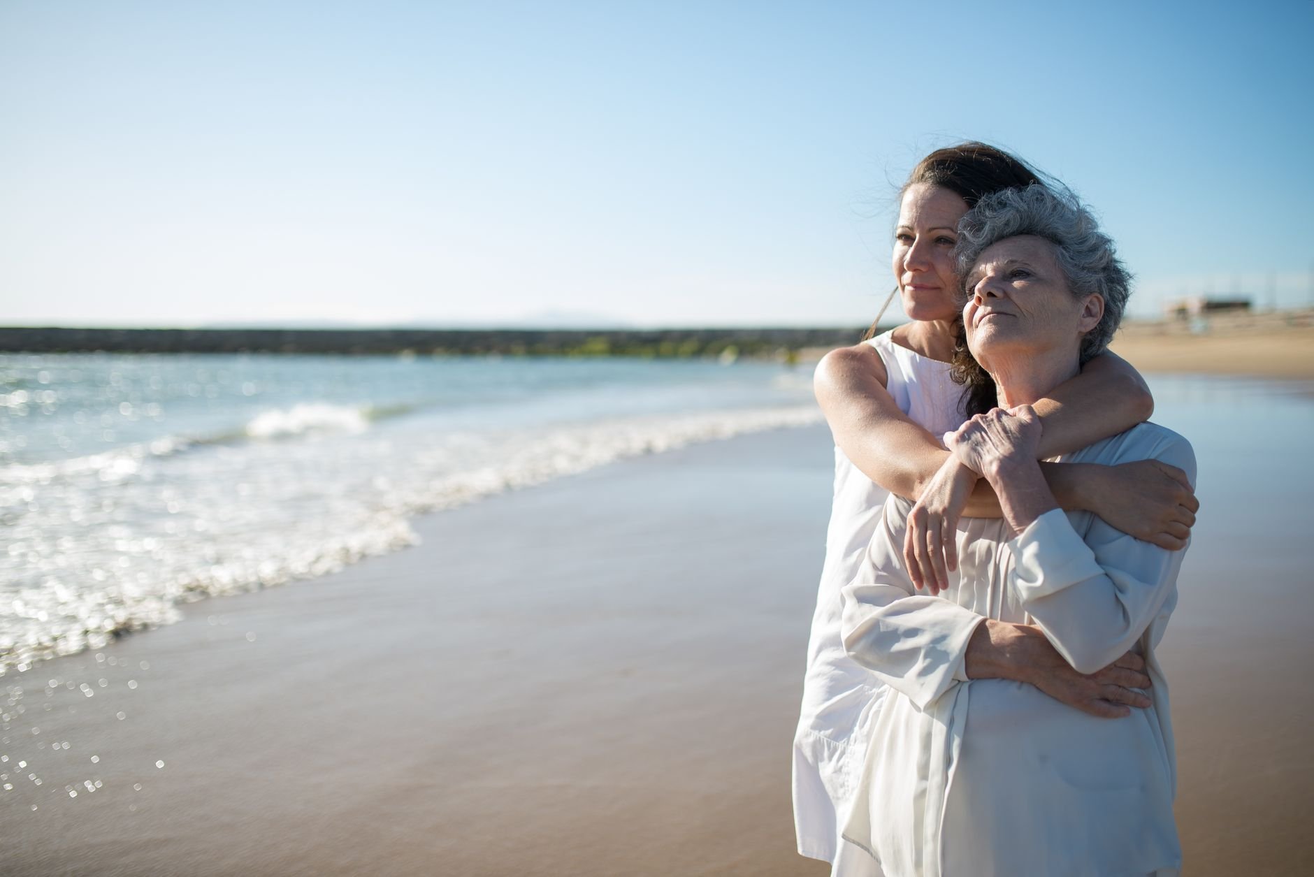 An elderly mother and adult daughter share a warm embrace on a sunny beach.